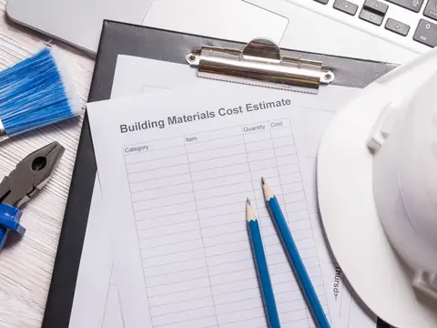 A blank cost estimate form with columns for category, item, quantity and cost on a clipboard, with two pencils, a hard hat, pliers and a paintbrush beside it