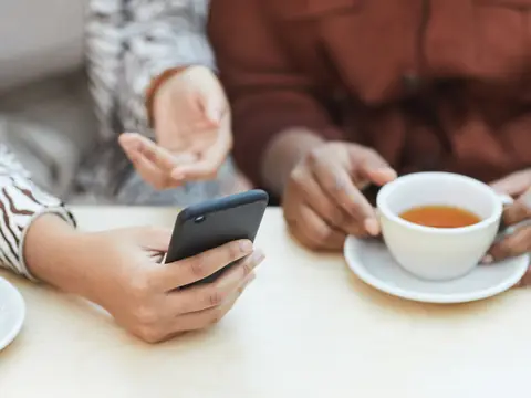 Two people at a table, one holding a smartphone and the other a cup of tea, seen from the shoulders down