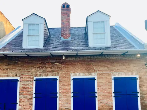 The slate roof of a brick Creole cottage in New Orleans, with two white dormers, a brick chimney with metal flashing at its base and metal-capped edges