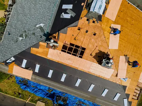 A roof seen from directly above during a re-roof: old shingles on one side, black underlayment, bare plywood decking with new sheets going down, and two workers at the edge