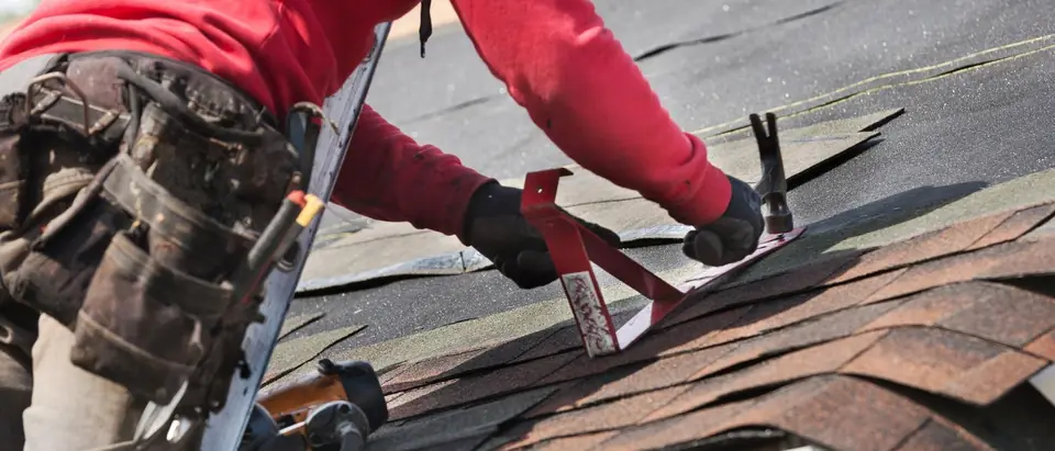 A roofer in a red sweatshirt and a worn tool belt, seen from behind, hammering a metal bracket onto asphalt shingles at the edge of a roof