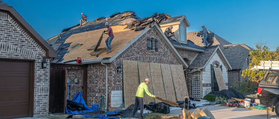 A crew stripping old shingles from the roof of a brick house, with bare decking showing, plywood leaning against the wall and tarps on the ground