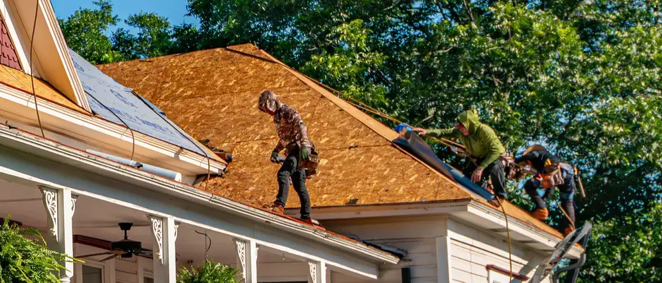 Three roofers on the bare wooden deck of a white frame house with a turned-post porch and hanging ferns, rolling out underlayment along a hip of the roof