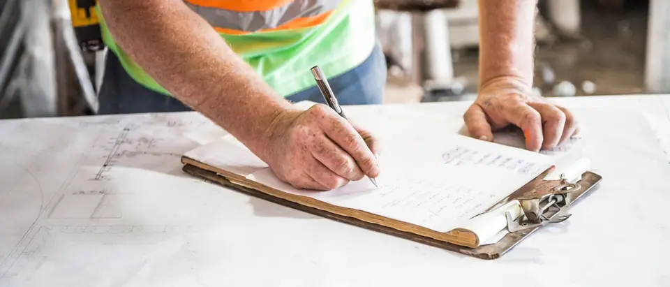 A worker in a high-visibility vest writing on a clipboard that lies on a set of building drawings