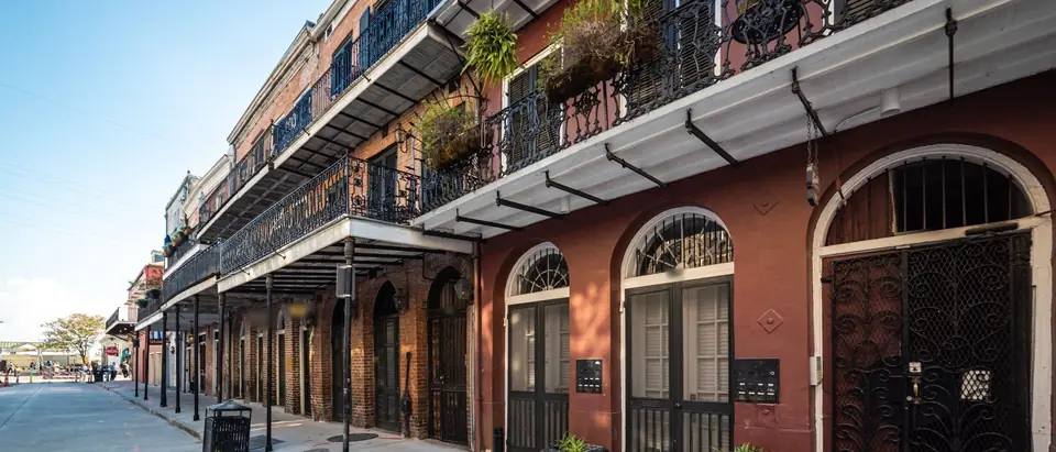 A row of old brick townhouses in the French Quarter of New Orleans with cast-iron galleries, arched doorways and hanging ferns along an empty sidewalk