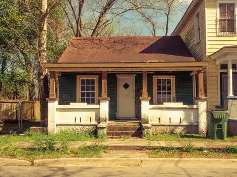 A narrow one-story wooden house with a hipped shingle roof and a deep front porch on brick piers, beside a taller two-story house