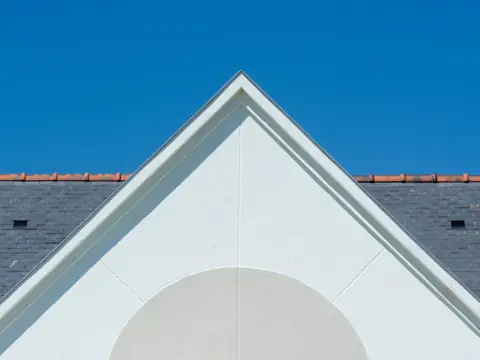 A grey slate roof topped with a row of terra cotta ridge tiles, behind a white gable against a blue sky