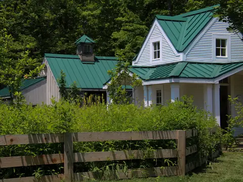 A white clapboard house and outbuilding with green standing seam metal roofs, behind a wooden rail fence and summer greenery